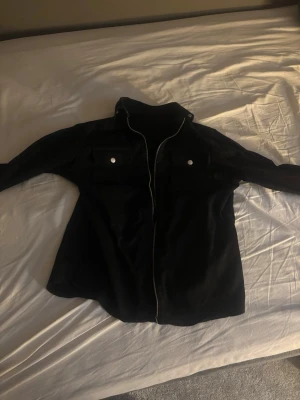 Svart overshirt med dragkedja - Stilren svart overshirt med dragkedja framtill och två bröstfickor med lock och silverfärgade knappar. Overshirten har klassisk krage och raka ärmar. Perfekt att slänga över en t-shirt för en avslappnad look.