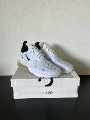 Nike Air Max 270 - Vit - Nike Air Max 270 sneakers i vitt med svarta detaljer och synlig Air-enhet i hälen. Ovandelen är i mesh för bra ventilation och har klassisk Nike-logga på sidan. 