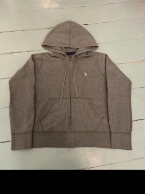 Grå zip hoodie från Polo Ralph Lauren - Klassisk grå hoodie med dragkedja från Polo Ralph Lauren. Har huva med dragsko, känguruficka och den ikoniska vita logon broderad på bröstet. Tillverkad i mjukt bomullsmaterial som är perfekt för en avslappnad stil.