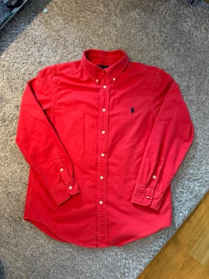 Röd skjorta från Ralph Lauren - Klassisk röd skjorta från Ralph Lauren med button-down krage och mörkblå broderad logga på bröstet. Skjortan har långa ärmar med vita knappar. Perfekt för en stilren och avslappnad look.