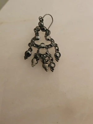 Svart hängande örhänge med stenar - Unikt svart örhänge i metall med flera gnistrande svarta stenar och små hängande detaljer. Örhänget har en bohemisk, vintageinspirerad design med runda former och dekorativa element som ger en cool och personlig stil. Ej ett par. Tappat bort det ena. Har aldrig användts.  