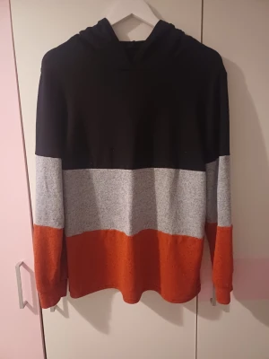 Hoodie  - Snygg hoodie med färgblock i svart upptill, grått i mitten och orange nedtill samt på ärmarna. Mjuk och bekväm med huva, perfekt för chill dagar. Ingen synlig logga eller tryck. Materialet känns som bomullsblandning.