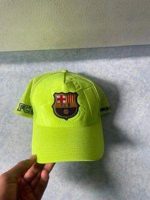 Neongul FC Barcelona keps - Snygg neongul keps från FC Barcelona med klubbens emblem broderat framtill. Kepsen har mesh på sidorna och bak för extra ventilation samt justerbar snapback. Texten 'FCBARCELONA' och '1899' finns broderad på baksidan och sidan. Perfekt för dig som älskar fotboll och vill sticka ut.