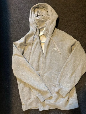 Grå hoodie från Nike med dragkedja - Klassisk grå hoodie från Nike med vit logga på bröstet. Hoodien har hel dragkedja, snörning i huvan och två stora fickor framtill. Mjuk insida i fleece för extra komfort. Perfekt för en avslappnad och sportig stil.