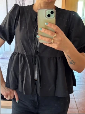 Svart volangblus med knytband - Snygg svart blus med volanger och knytband framtill. Blusen har rund halsringning och korta puffärmar. Tillverkad i ett lätt tyg som ger ett luftigt intryck. Perfekt att styla med jeans eller kjol för en trendig look. ALDRIG ANVÄND‼️