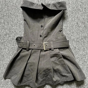 Grå axelbandslös kort klänning - Trendig grå axelbandslös kort klänning med tunna vita ränder, knappar framtill och ett brett bälte i midjan. Klänningen har veckad kjol med inbyggda shorts.