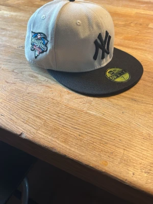 New Era Yankees keps - Snygg 59FIFTY keps från New Era med New York Yankees-logga i svart framtill och World Series 1999 patch på sidan. Kepsen är grå med svart skärm och grön undersida. Klassisk flatbrim-modell i bomull, perfekt för dig som gillar streetwear och sportig stil.