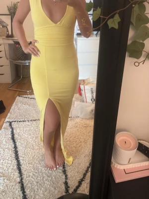 Gul långklänning med slits - Snygg gul långklänning med djup v-ringning och markerad midja. Klänningen är ärmlös och har en hög slits framtill som ger en trendig look. Perfekt för dig som vill sticka ut med en färgstark och stilren outfit.