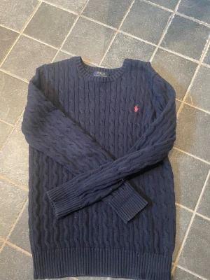 Mörkblå kabelstickad tröja Polo Ralph Lauren - Kabelstickad mörkblå tröja från Polo Ralph Lauren med rund halsringning och klassisk röd logga broderad på bröstet. Tröjan har långa ärmar och ribbade muddar vid ärmslut och nederkant. Perfekt för lager på lager och snygg till jeans.