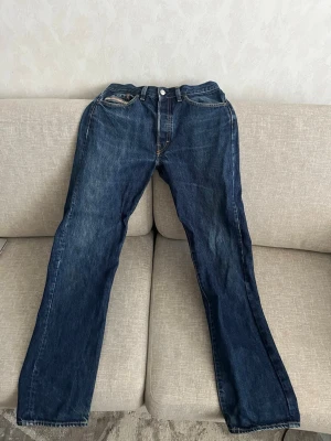 Diesel Industry blå raka jeans - Klassiska blå jeans från diesel industry med rak passform och fem fickor. Jeansen har tydliga kontrastsömmar och en brun läderpatch bak i midjan. Tillverkade i slitstark denim, perfekta för en avslappnad och tidlös stil.