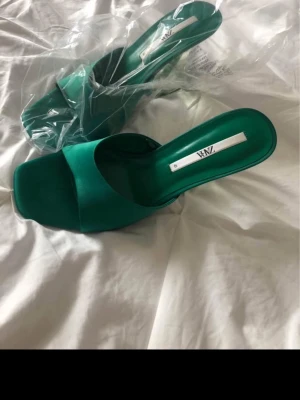 Gröna slip-in klackar från Zara - Snygga gröna slip-in klackar från Zara med bred rem över foten och fyrkantig tå. Klacken är genomskinlig och ger en modern touch. Ovandelen är i textil och sulan i gummi. Perfekta för dig som vill sticka ut med färg och stil.