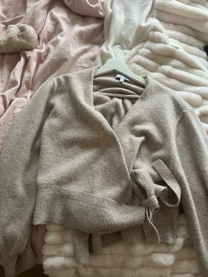 kofta & Other Stories - Mysig beige stickad omlottkofta från & Other Stories. Koftan har långa ärmar, bred ribbad mudd vid ärmslut och midja samt knytband i sidan. Perfekt för lager på lager och en avslappnad men snygg look.