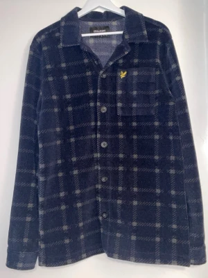 Mörkblå rutig overshirt Lyle & Scott - Overshirt i mjukt material från Lyle & Scott med mörkblå och grå rutig design. Klassisk krage, knappstängning framtill och bröstficka med gul logga. Perfekt lager på lager-plagg för en avslappnad stil.