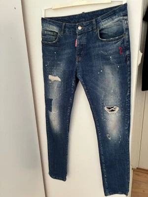 Dsquared2 blå slitna jeans - Säljer ett par blå jeans från Dsquared2 med distressed detaljer, hål och färgstänk för en cool streetwear-look. Klassisk femficksmodell med röd logotyppatch bak. Jeansen har normal passform och är tillverkade i denim.