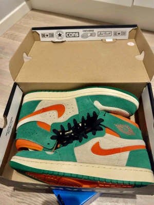 Nike Air Jordan 1 Mid grön/orange - Nike Air Jordan 1 Mid sneakers i grön mocka med orange detaljer och swoosh, beige paneler och svart snörning. Klassisk Air Jordan-logga på sidan och vit mellansula. Skön retrostil med färgstark look och slitstark yttersula.