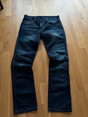 Mörkblå raka jeans från Levi's - Säljer ett par mörkblå jeans från Levi's med klassisk rak passform och fem fickor. Jeansen har snygga slitningar framtill och bakfickor med den ikoniska Levi's-sömmen. Tillverkade i slitstarkt denimtyg med knappgylf.