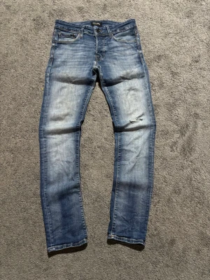 Jack & Jones jeans  - Säljer ett par blå jeans från Dr. Denim med snyggt slitna detaljer och en liten reva på ena benet. Jeansen har klassisk femficksmodell, normal midja och smal passform. Tillverkade i mjuk denim med tvättad look.