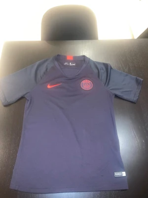 Mörkblå PSG t-shirt från Nike - Snygg mörkblå t-shirt från Nike med PSG-logga och röd Nike-swoosh på bröstet. Tillverkad i Dri-FIT-material som andas, perfekt för träning eller sport. Kortärmad modell med rund hals och diskreta röda detaljer. Den är liten i storlek