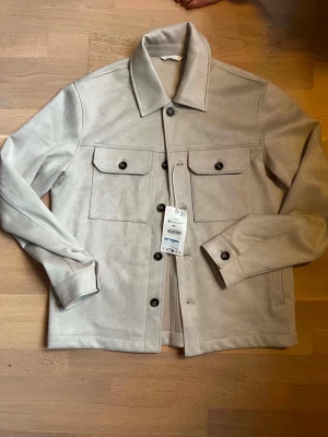 Beige overshirt från ZARA - Stilren beige overshirt från ZARA med klassisk krage, två stora bröstfickor med lock och svarta knappar. Tillverkad i ett mjukt material som ger en lyxig känsla. Perfekt att bära öppen eller stängd för en avslappnad look.
