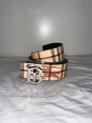 Beige och brunt rutigt Burberry-bälte - Snyggt bälte från Burberry i beige och brunt med klassiskt rutigt mönster och detaljer i form av Burberrys logga i metall. Bältet har en bred design och är tillverkat i skinn med ett exklusivt intryck. Perfekt accessoar för att lyfta din outfit.