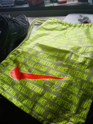 Neongul gympåse från Nike - Snygg neongul gympåse från Nike med dragskostängning och tryck i flera språk. Stor röd Nike-logga på framsidan och text som 'NIKE VOETBAL', 'NIKE FUTEBOL' och 'NIKE CALCIO'. Tillverkad i lätt syntetmaterial, perfekt för träning eller skola.