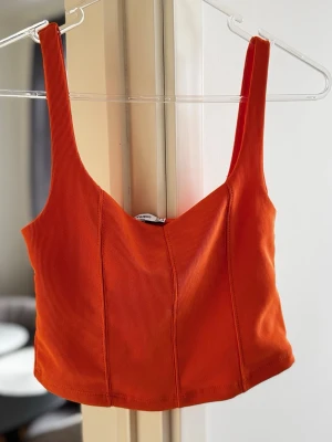 Ribbad orange crop top - Säljer en ribbad orange crop top med breda axelband och figurnära passform. Toppen har en enkel men snygg design som passar perfekt till sommaren. Materialet är stretchigt och bekvämt.