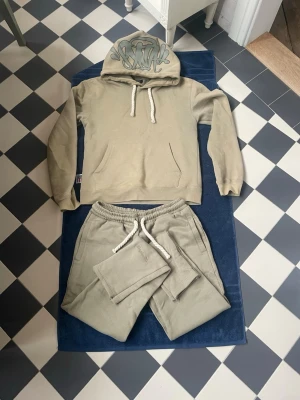 Olivgrönt set med hoodie och byxor från ”SYNA” - Säljer ett olivgrönt set från ”SYNA” med hoodie och shorts i mjukt material. Hoodien har vita drag snören och coolt graffiti-tryck på huvan. Byxorna har matchande tryck bak och elastisk midja med vita dragsnören. Perfekt för en avslappnad och trendig look. Påsen ingår i detta priset. 
