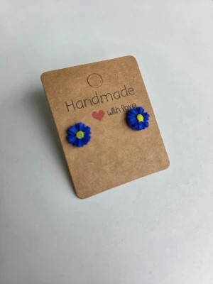 Blå blomformade handgjorda örhängen  - Supersöta handgjorda örhängen i form av blå mini prästkragar med gul mitt. Perfekta till midsommar! Örhängena är små och diskreta, tillverkade i polymerlera och sitter på stift av rostfritt stål. 