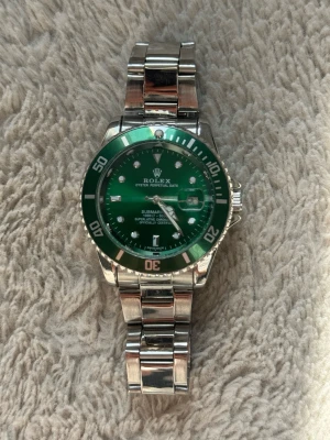 Rolle Submariner grön/silver klocka - Snygg Rolle Submariner med grön urtavla och grön vridring. Klockan har en robust länk i silverfärgat stål och tydliga indexmarkeringar. Klassisk design med datumfönster och ikonisk logga på urtavlan. Perfekt för dig som gillar exklusiva accessoarer.