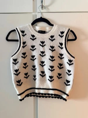 Vit stickad väst från New Look - Supersöt vit stickad väst från New Look med svart blommigt mönster. Rund halsringning och ribbade kanter. Perfekt att styla över en skjorta eller t-shirt.