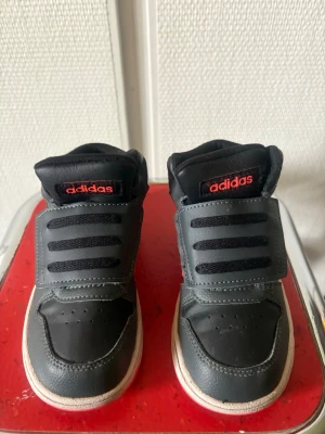Svarta Adidas sneakers med röda detaljer för barn 👧  - Snygga svarta sneakers från Adidas med röda detaljer och klassiska tre ränder på sidan. Skorna har kardborreband och snörning, samt en vit sula. Ovandelen är i syntetmaterial och mesh för extra komfort. Perfekta för dig som gillar sportig stil.