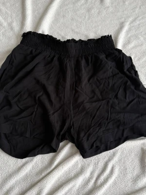Svarta loose shorts med resårmidja (S) - Säljer ett par svarta shorts med bred resår i midjan och avslappnad passform. Shortsen har fickor på sidorna och är gjorda i ett mjukt, tunt tyg som passar perfekt till varma dagar. Stilrena och enkla att matcha med olika toppar.