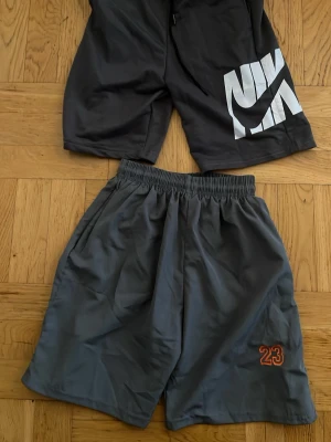 Två par shorts en nike en 23 - Två par svarta shorts från Nike. Ena paret har stor vit Nike-logga på benet och det andra har en liten orange '23' broderad på benet. Båda har elastisk midja och är tillverkade i ett lätt, syntetiskt material. Perfekta för sport eller chill.