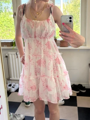 Vit och rosa blommig sommarklänning - Söt kortklänning i vitt med rosa blommönster och tunna axelband med knyt. Klänningen har volangdetaljer och en luftig, flowy passform som passar perfekt till varma dagar. Tillverkad i ett lätt och mjukt material, perfekt för sommarens alla festligheter 🌺💕☀️. Endast använd vid en skolavslutning!