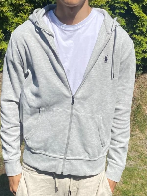 Vit hoodie från Polo Ralph Lauren - Snygg ljusgrå hoodie från Polo Ralph Lauren med dragkedja, huva med dragsko och känguruficka. Klassisk logga broderad på bröstet. Tillverkad i mjukt bomullsmaterial som känns skönt mot huden. Perfekt för en avslappnad och stilren look.