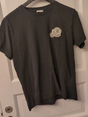 Svart Moncler t-shirt med patch - Snygg svart t-shirt från Moncler med klassisk rund hals och korta ärmar. På bröstet sitter en stor broderad Moncler-patch i vitt och blått som ger en exklusiv känsla. Tillverkad i mjuk bomull som känns skön mot huden. Perfekt till jeans eller shorts.