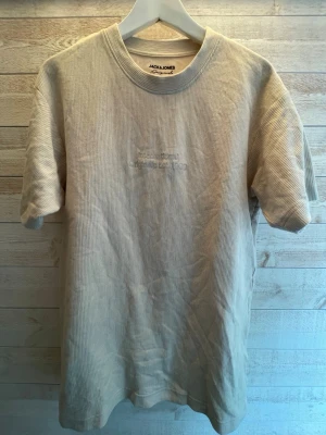 Beige ribbad t-shirt från Jack & Jones - Säljer en beige ribbad t-shirt från Jack & Jones med diskret broderad logga på bröstet. T-shirten har rund halsringning och korta ärmar. Perfekt för en avslappnad och stilren look.