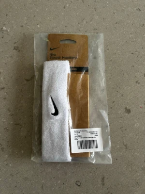 Nike vit pannband med Swoosh-logga - Nike Swoosh Headband i vitt med klassisk svart Swoosh-logga broderad framtill. Tillverkad i mjukt och stretchigt material som sitter bekvämt runt huvudet. Perfekt för att hålla håret på plats under träning eller till vardagslooken.
