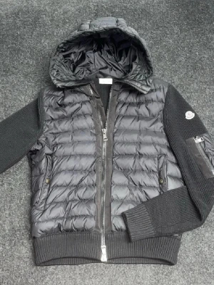 Moncler cardigan - Svart moncler cardigan storlek L med stickade ärmar och quiltad kropp. Jackan har huva, dragkedja framtill och ribbade muddar. På ena ärmen finns en ficka med dragkedja och Moncler-logga. Perfekt för dig som gillar streetstyle och vill ha något som sticker ut. Svarar alla meddelanden direkt och fraktar alltid samma dag som köp:)