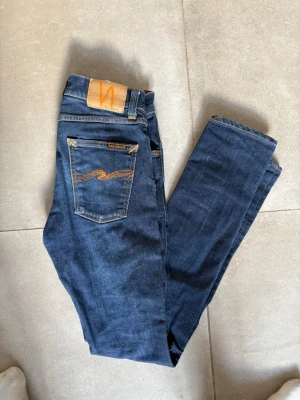 Nudie jeans  - Säljer ett par mörkblå jeans från Nudie Jeans med klassisk femficksmodell och snygga kontrastsömmar. Jeansen har raka ben och en patch i läder baktill med Nudie-loggan. Materialet är mjukt denim i bomull och passformen är slim. W27 L32