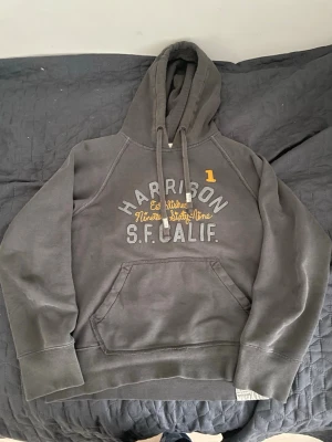 Mörkgrå hoodie Harrison S.F. Calif. - Mörkgrå hoodie från Harrison med tryck i vitt och gult på bröstet där det står 'Harrison S.F. Calif.' och 'Established Nineteen Sixty-Nine'. Klassisk känguruficka framtill, huva med dragsko och ribbade muddar. Perfekt för en avslappnad streetwear-look. Storlek M men sitter snarare som en S