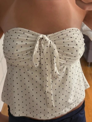 Prickig  tubtopp - Söt vit tubtopp med svarta prickar och knyt framtill. Toppen är axelbandslös och har en avslappnad passform nedtill. Perfekt för varma dagar och enkel att styla med jeans eller kjol. 