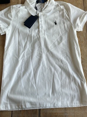 Vit pikétröja från Ralph Lauren - Klassisk vit pikétröja från Ralph Lauren med korta ärmar och krage. Tröjan har knappar framtill och den ikoniska broderade loggan på bröstet. Perfekt till jeans eller kjol för en clean och stilren look.