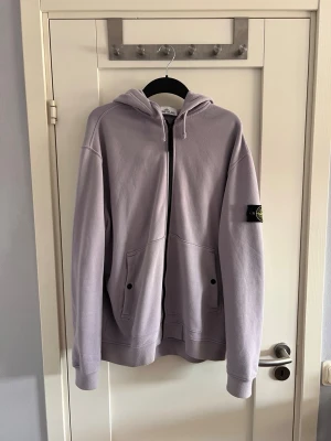 Ljuslila hoodie från Stone Island - Säljer en ljuslila hoodie från Stone Island med dragkedja framtill och två stora fickor. Klassisk patch med logga på vänster ärm. Hoodien har huva med dragsko och ribbade muddar. Perfekt för dig som gillar streetwear och vill ha något stilrent.