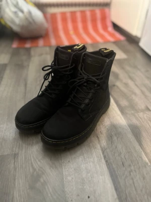 Svarta kängor från Dr. Martens - Säljer ett par svarta kängor från Dr. Martens med klassisk gul söm och högt skaft. Kängorna har snörning hela vägen upp och är tillverkade i slitstark textil med detaljer i läder. Ikonisk flärp med gul text baktill.