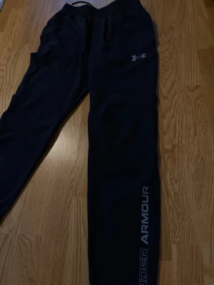 Svarta träningsbyxor Under Armour - Snygga svarta träningsbyxor från Under Armour med logga på låret och text längs benet. Byxorna har resår i midjan och vid bensluten, samt dragsko för justerbar passform. Perfekta för gymmet eller löprundan.