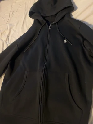 Svart hoodie från Polo Ralph Lauren - Klassisk svart hoodie med dragkedja från Polo Ralph Lauren. Hoodien har huva med dragsko, två fickor framtill och den ikoniska vita broderade loggan på bröstet. Perfekt för en chill och stilren look.