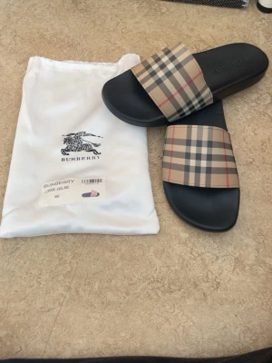 Burberry toflor med klassiskt rutmönster - Snygga sandaler från Burberry med svart sula och bred rem i det ikoniska beige, röda och svarta rutmönstret. Märket är präglat på sulan och sandalerna har en modern, clean look. Perfekta för sommaren och ger en lyxig touch till din outfit.