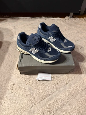 New Balance 2002r skor - Säljer ett par stilrena New Balance 2002r sneakers i mörkblått med vita och grå detaljer. Skorna har klassisk snörning och är tillverkade i en mix av mocka och mesh. Kom pm vid frågor?