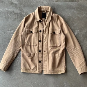 J.Lindeberg Overshirt / Utility-jacka – Beige (Strl S) - Beige jacka/overshirt i kraftigare bomullskvalitet från J.Lindeberg. Modellen stängs med mörka knappar framtill och är utrustad med fyra framfickor: två bröstfickor med knapplock samt två öppna insticksfickor nertill. Manschetter med knäppning vid ärmsluten.